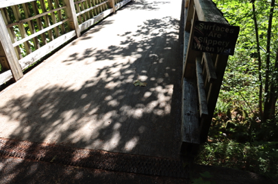 Paved surface transitions to wooden footbridge with railings over river – slippery when wet – slight lip at transitions